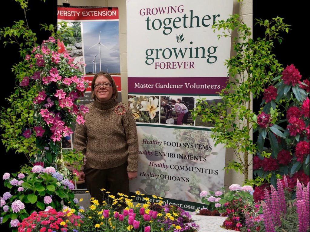 Jenny Crowe stands in front of a sign that says "Growing together, growing forever" with colorful flowers and plants surrounding her.