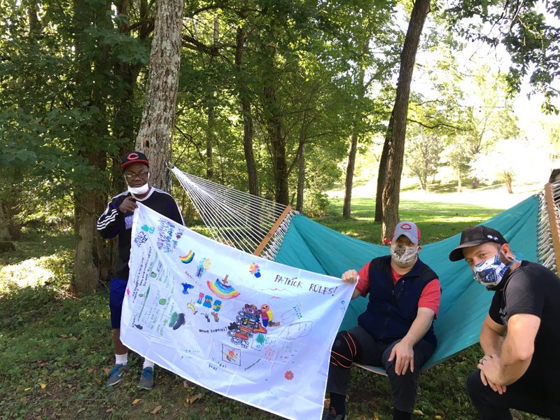 Photo of three men outside together with a hammock behind them. Two are holding a flag together.