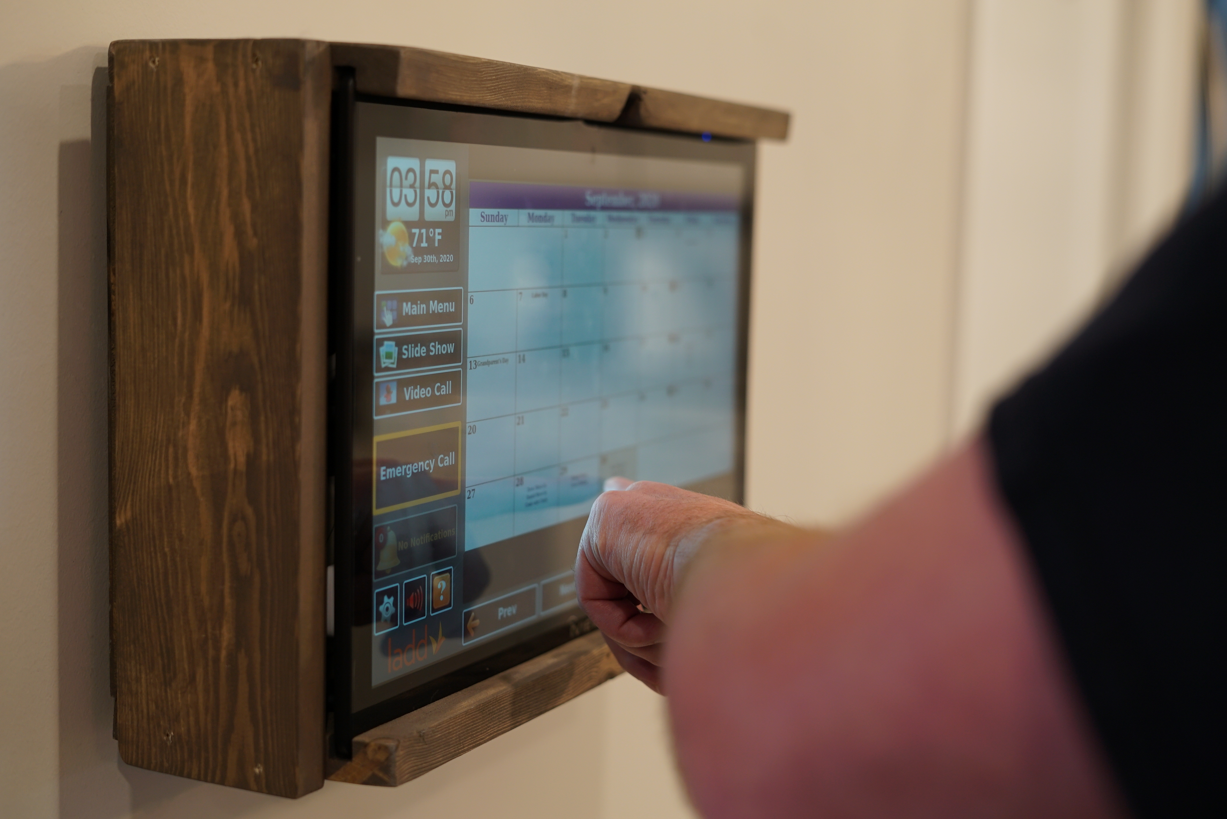 Someone is touching a virtual screen mounted to a wall. The virtual screen shows things like a calendar, emergency call button, schedule button and more.