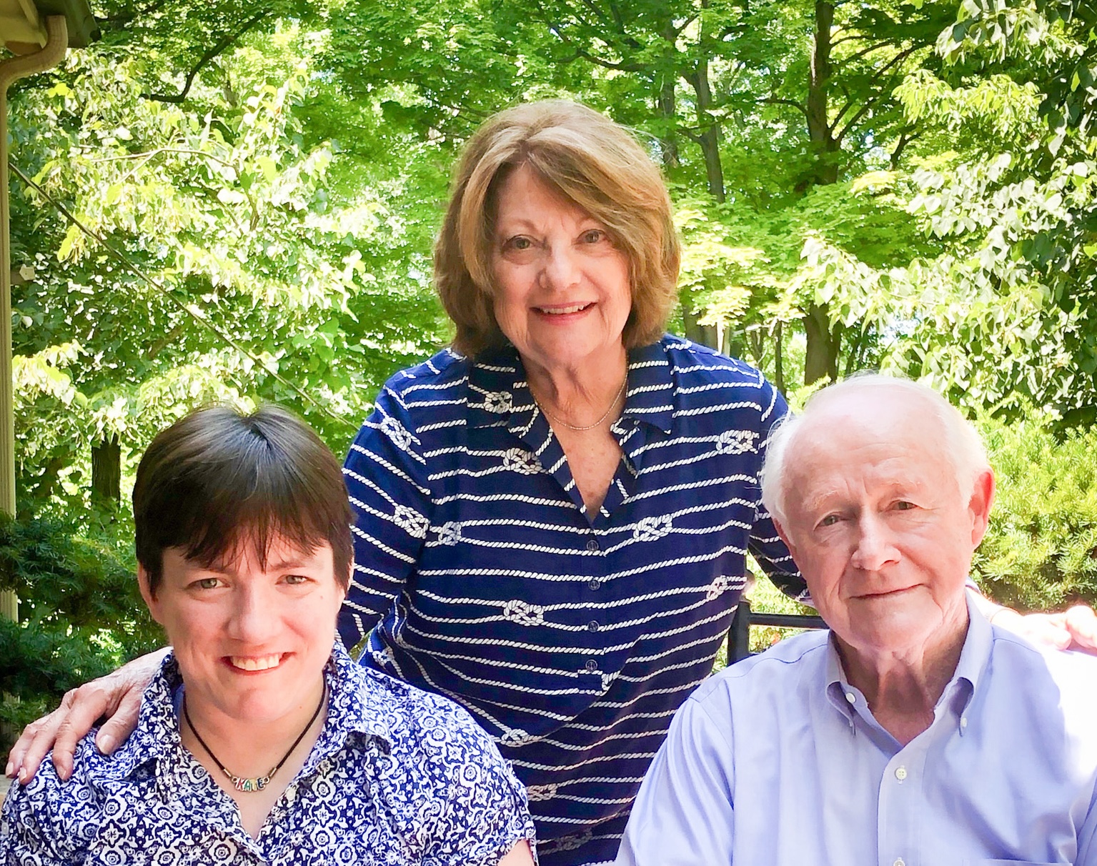 Family photo of Phyllis Breen standing between her daughter on the left and husband on the right with green foliage and sunlight in the backgroud.