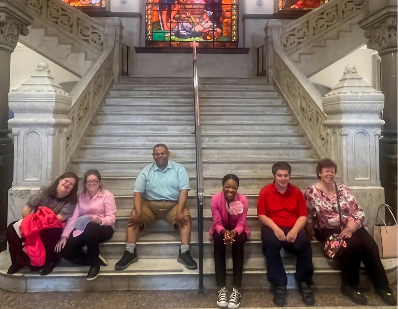 Six individuals sit on a grand marble staircase inside a historic building with ornate railings and stained glass windows. The group, diverse in age and appearance, includes three women and three men. They are smiling and appear to be enjoying their time together. The setting is elegant, with intricate architectural details and a warm, welcoming atmosphere.